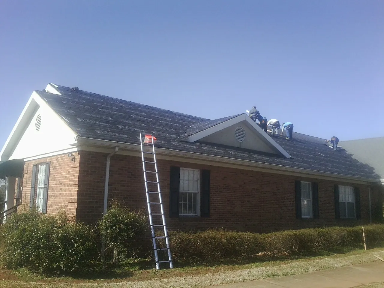 Crew tearing shingles off a brick ranch roof with a ladder set — Emergency Roof Repair work by our crew in Icard