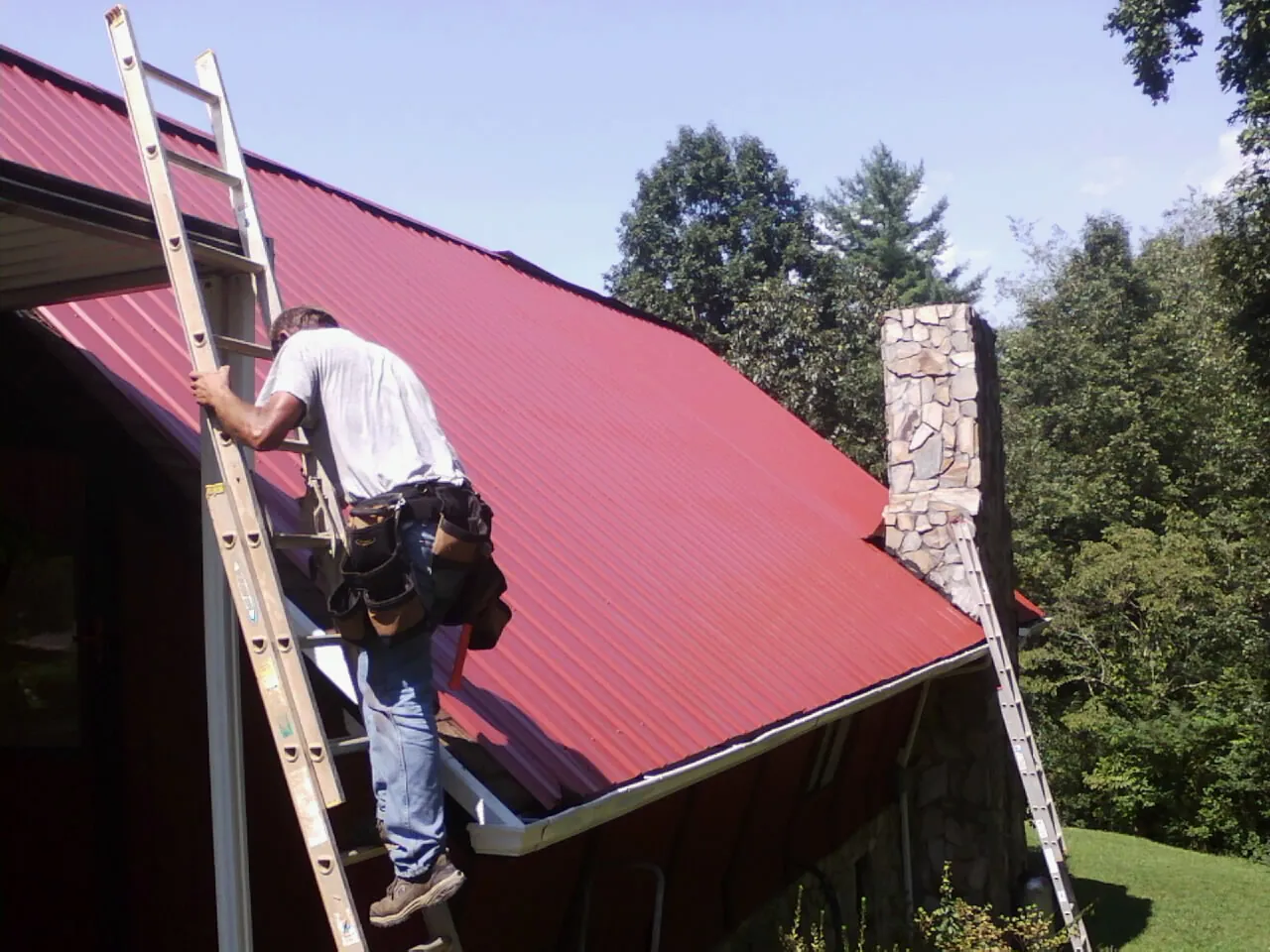Roofer on a ladder beside a red metal roof and stone chimney — Soffit & Fascia Repair by Roofing Brothers in Icard